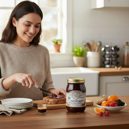 Woman spreading Broke Man's Pantry Gator Jam on toast in a bright kitchen with fresh fruit and ginger on the table.